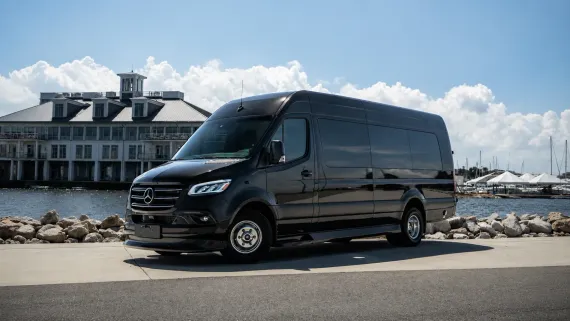 A black Mercedes-Benz Sprinter van parked on an asphalt lot near a building by the water under a clear blue sky.