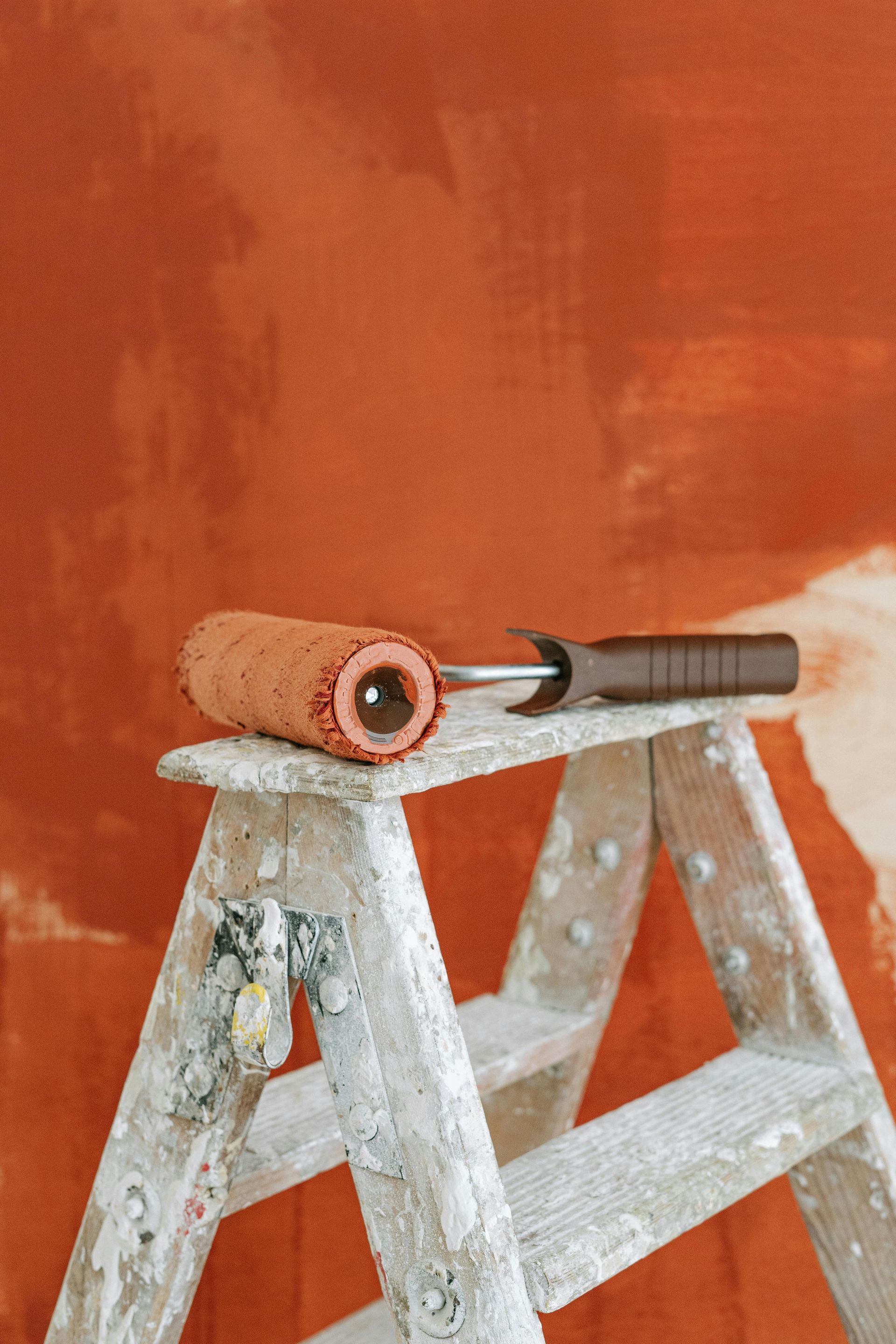 Paint roller and brush resting on a paint-splattered stepladder against an orange wall