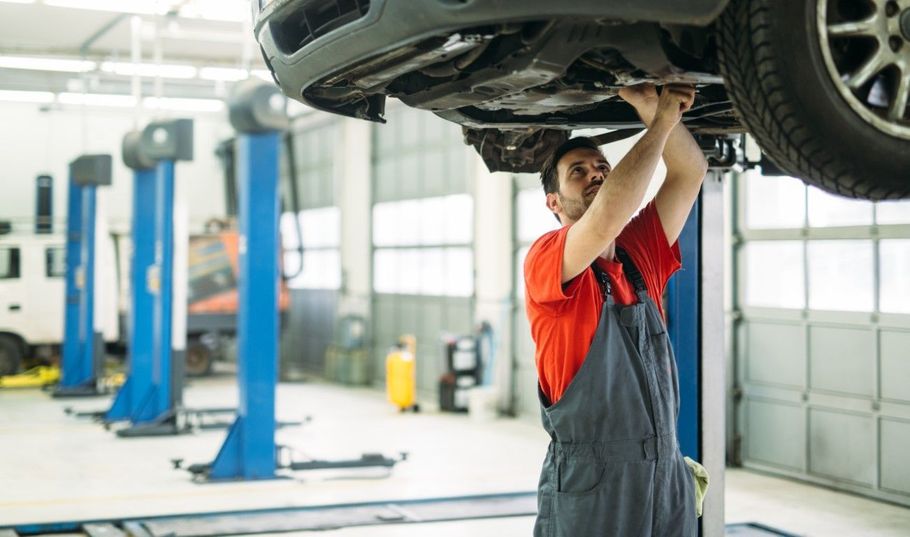 Mechanic in red shirt and overalls working under a raised car in an auto repair shop