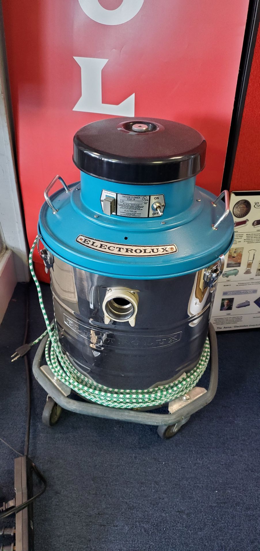 Blue cylindrical industrial vacuum cleaner on wheels with a black top, in front of a red wall.