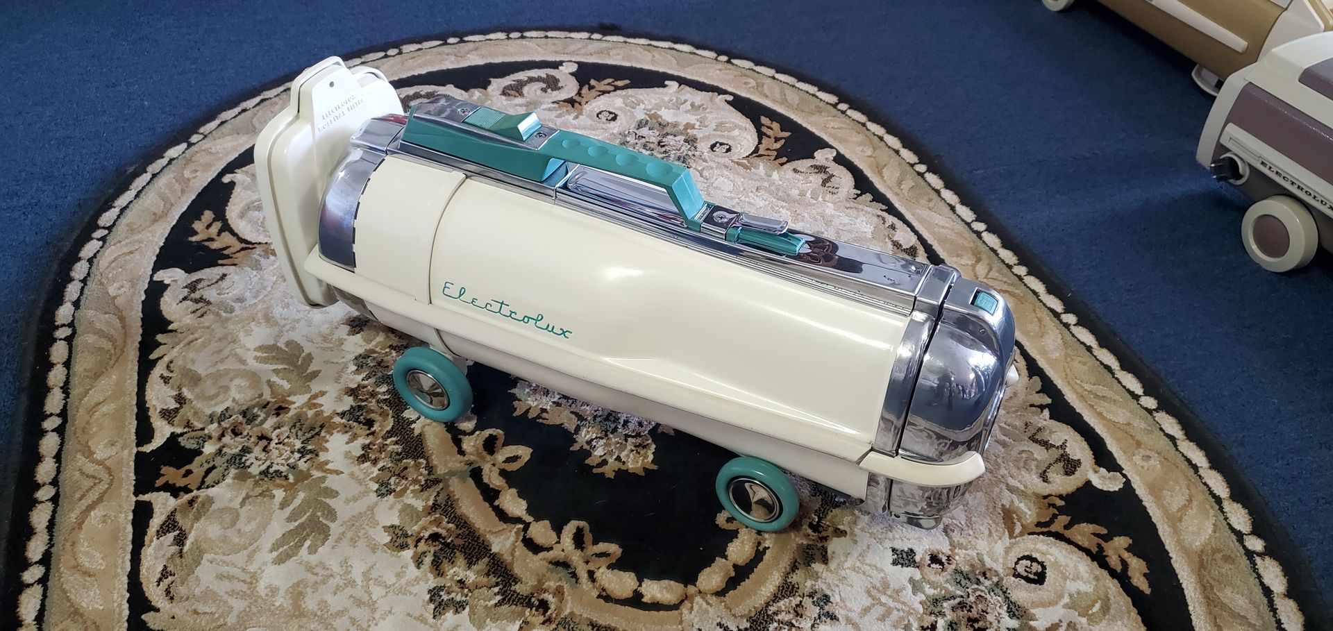 White cylindrical robot on blue carpet, with small wheels and a clear front, displayed on an ornate rug.
