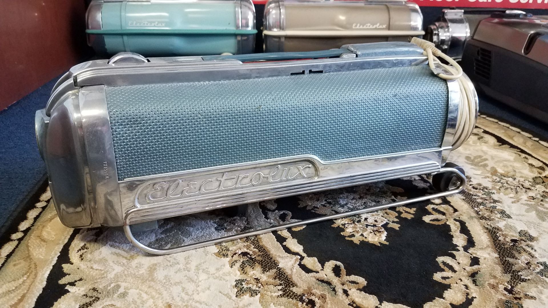 Blue cylindrical luggage case on a patterned rug, with other cases stacked in the background.