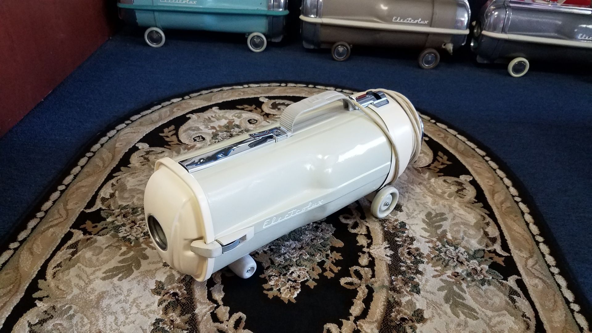 White cylindrical vacuum lying on a patterned rug in a room with toy trucks in the background