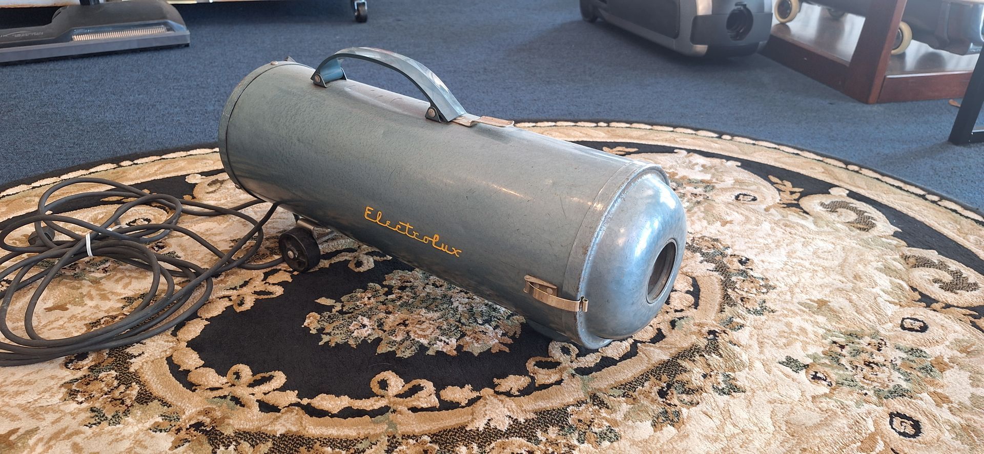 Gray cylindrical portable heater on an ornate rug in a room, with a handle on top