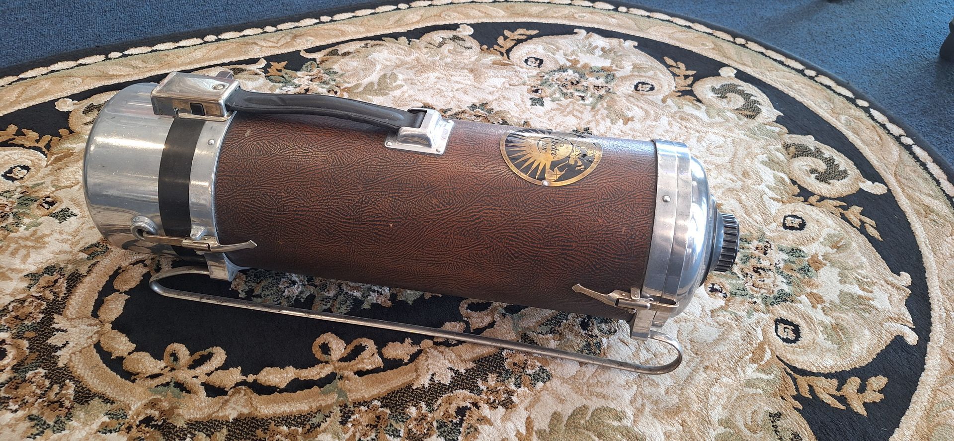 Brown cylindrical upholstered ottoman with silver ends on an ornate cream and black rug