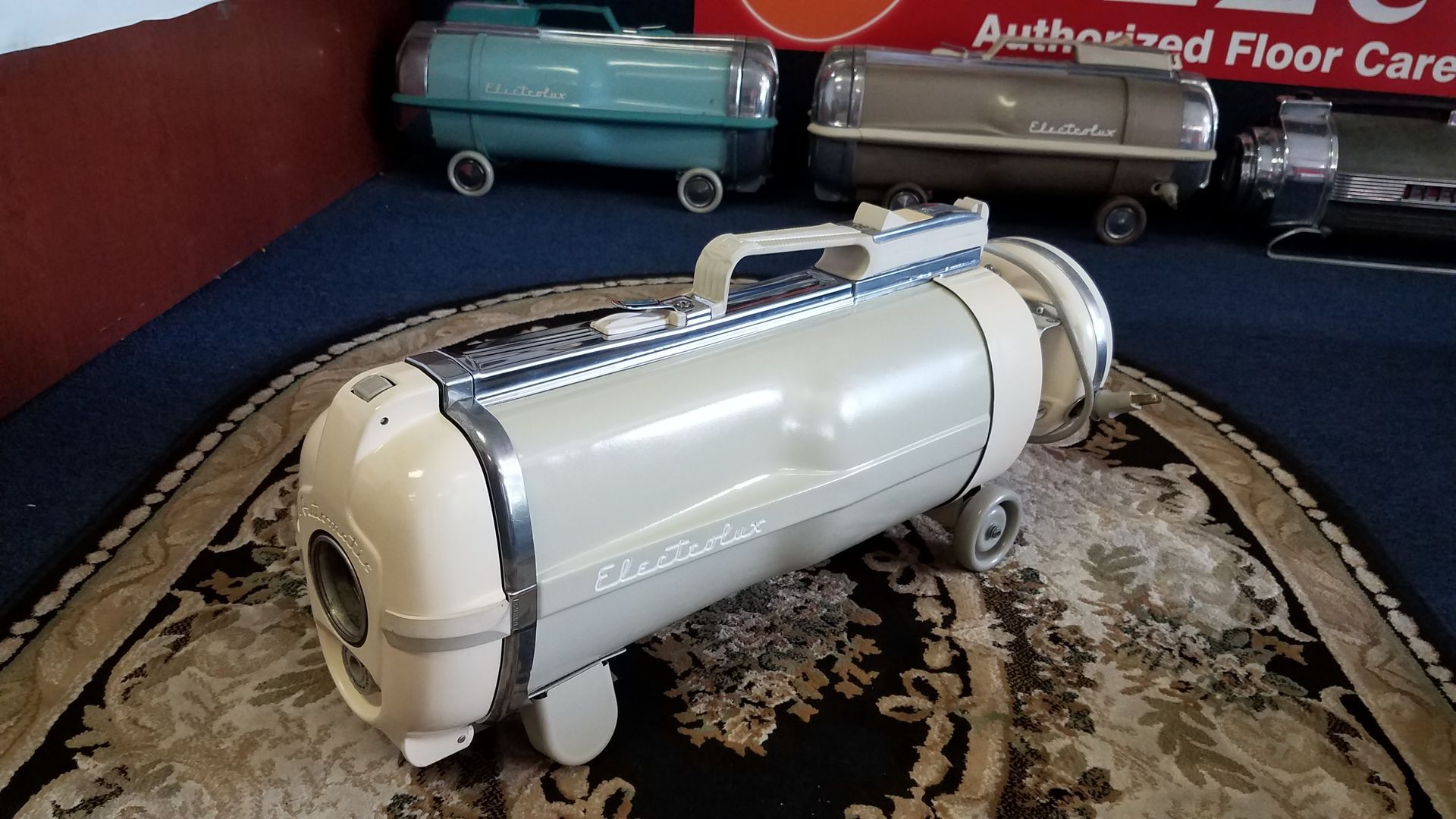 White cylindrical tank-like model with black straps on a patterned rug, with a teal tank in the background