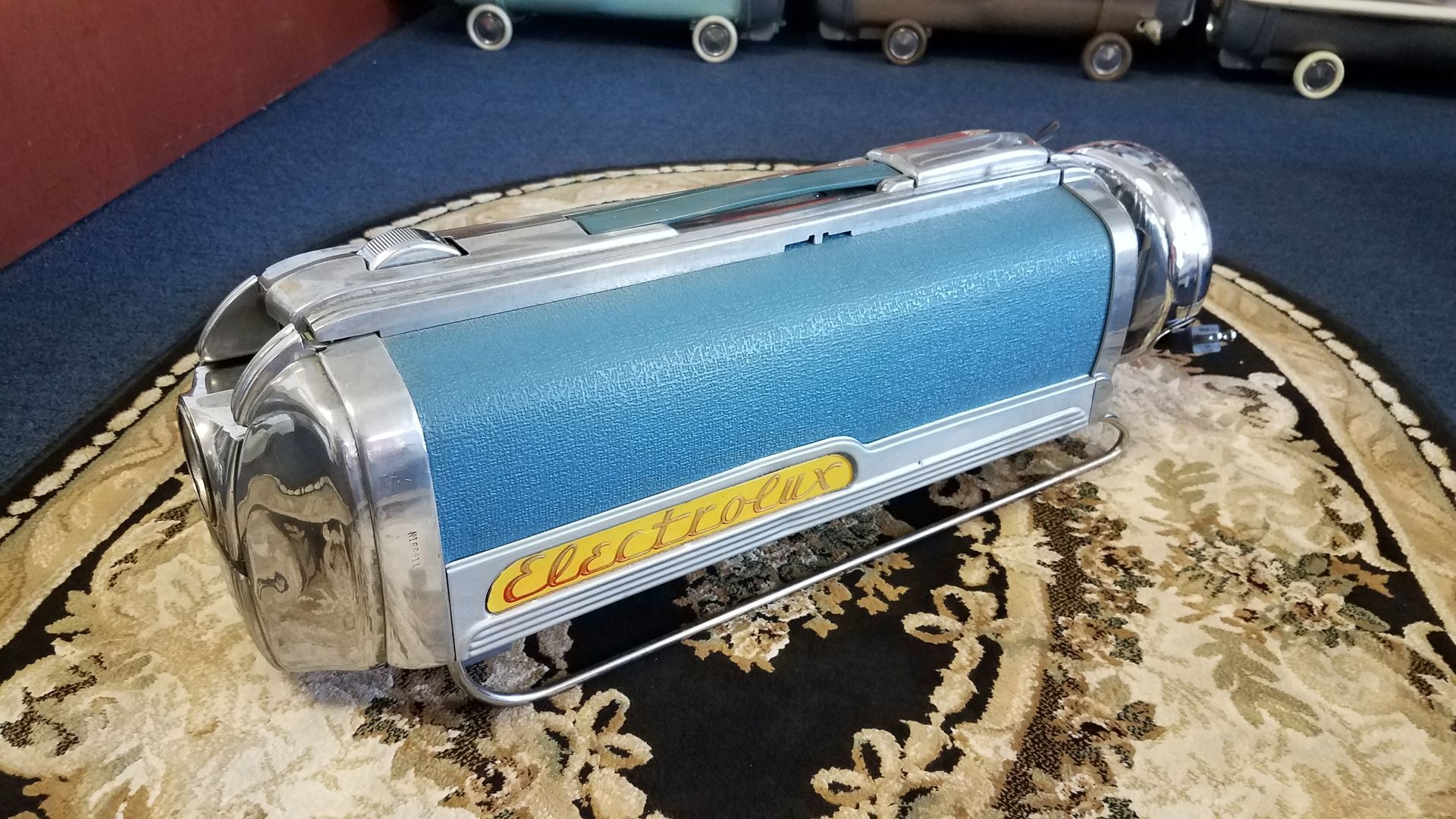 Blue cylindrical chrome audio speaker resting on a patterned tray top with an oval logo label