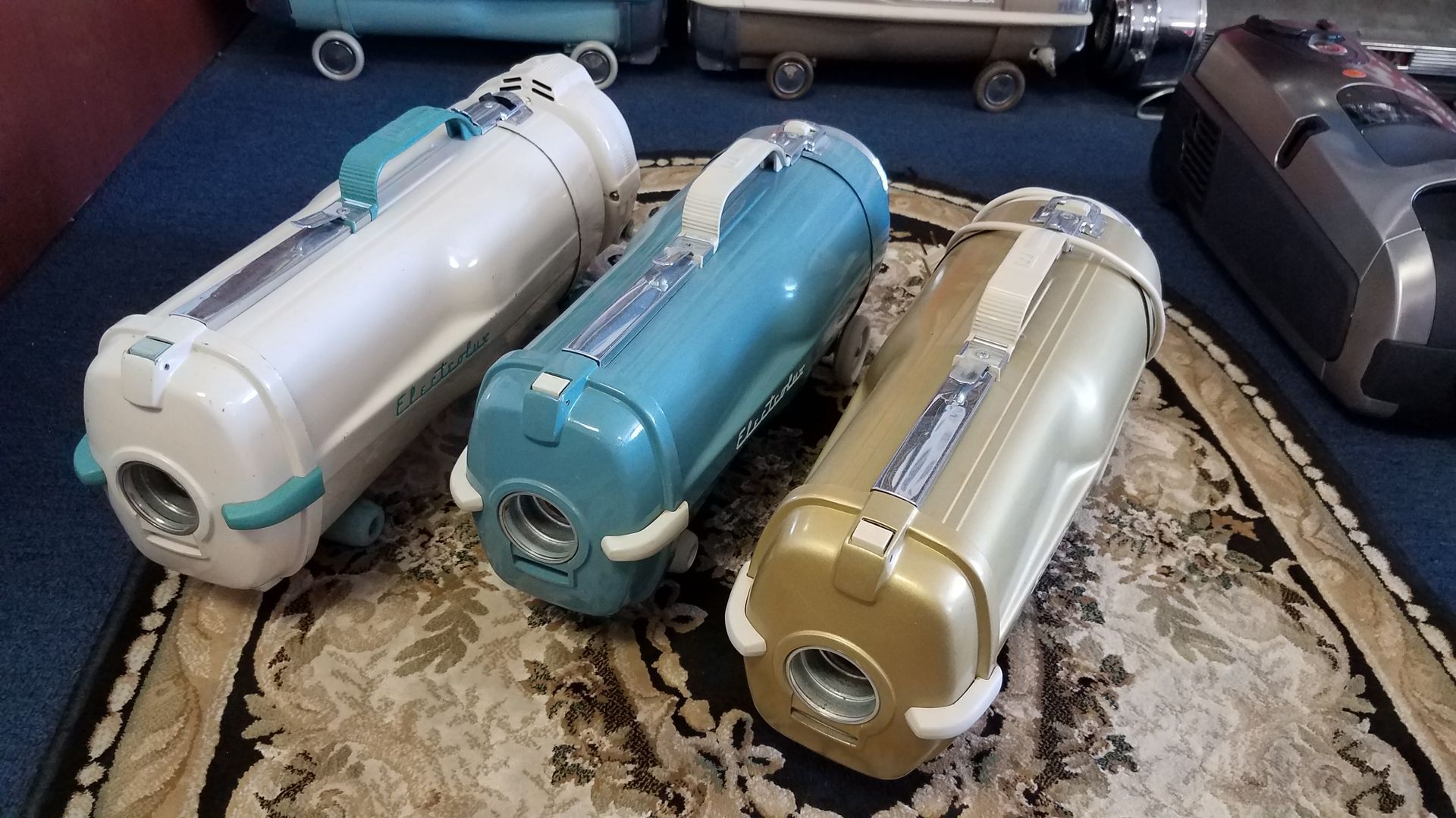 Three colorful vacuum cleaners lined up on a patterned rug, with a dark one partially visible at right.