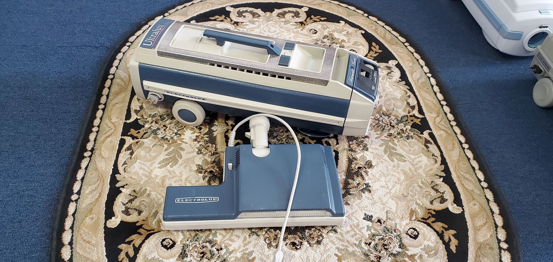 Blue-and-white toy van on a patterned rug beside a gray box and white cable, with a small card on top