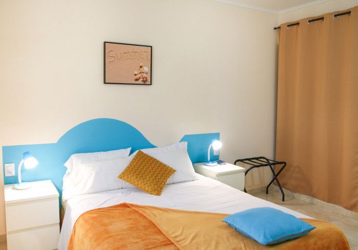 Bright bedroom with a white bed, blue pillows, orange blanket, bedside lamps, and beige curtains