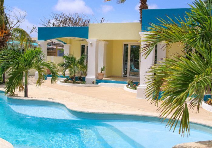 Tropical resort courtyard with a turquoise pool, cream villa, blue awnings, and palm trees