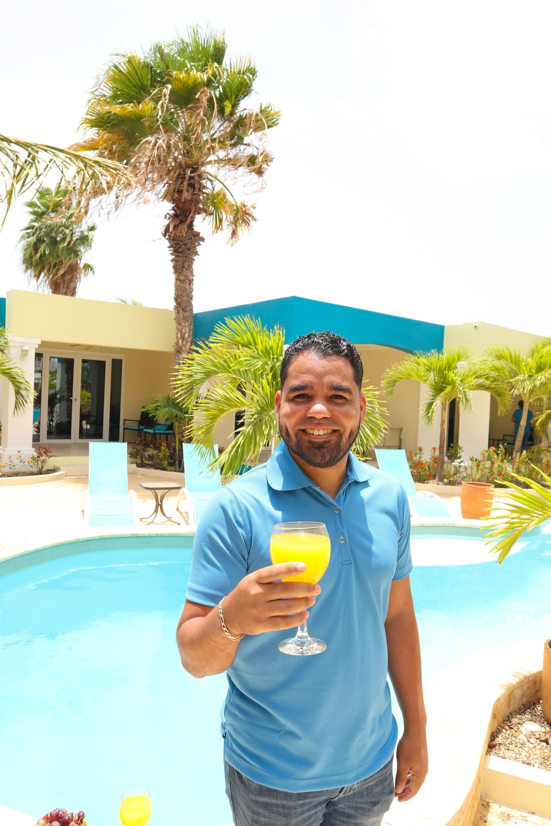 Man holding a yellow drink by a pool at a sunny resort with palm trees and lounge chairs
