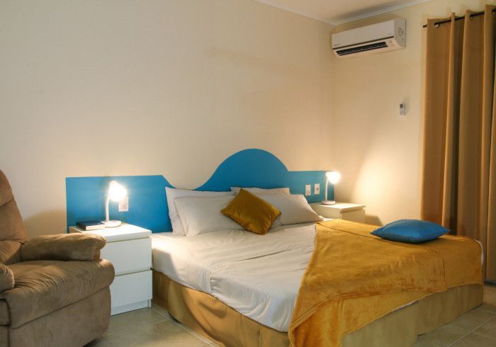 Neatly made bed in a beige bedroom with blue headboard, bedside lamps, armchair, and wall-mounted air conditioner