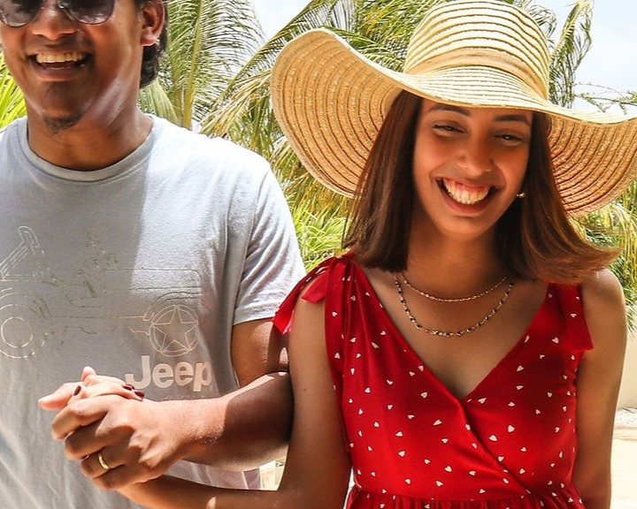 Smiling couple holding hands outdoors; woman wears a red dress and straw hat, man wears a gray T-shirt.