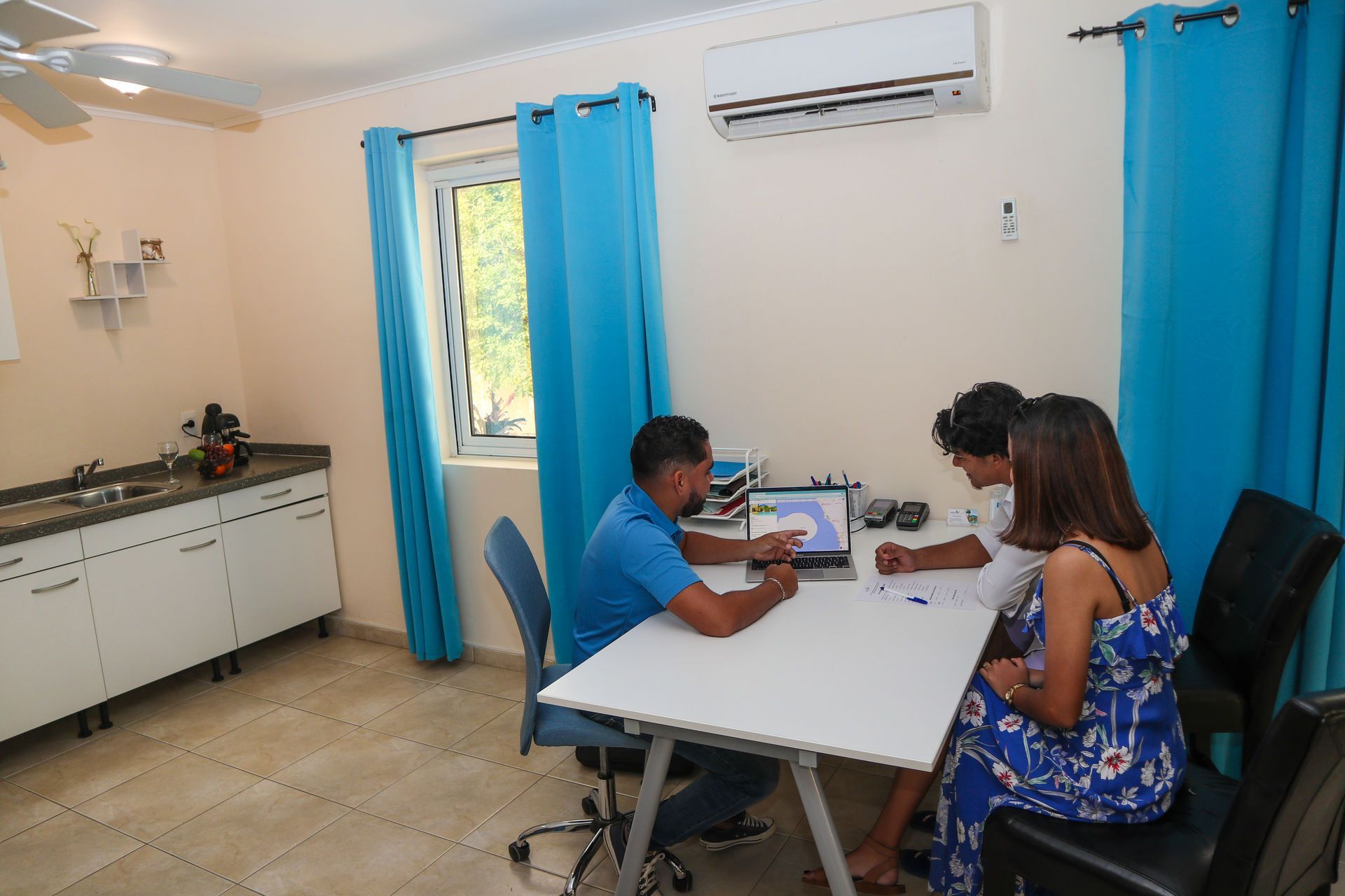 Two people sit at a table in a bright office, working on paperwork near blue curtains and a window.
