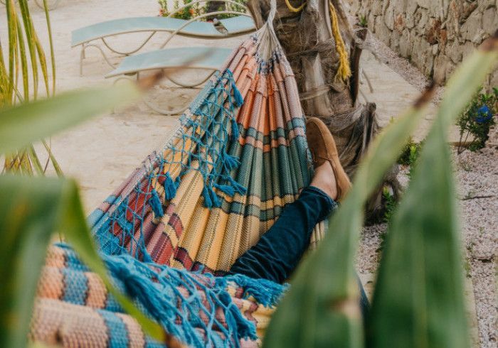 Colorful striped hammock beside a stone wall, with a person resting under leafy branches.