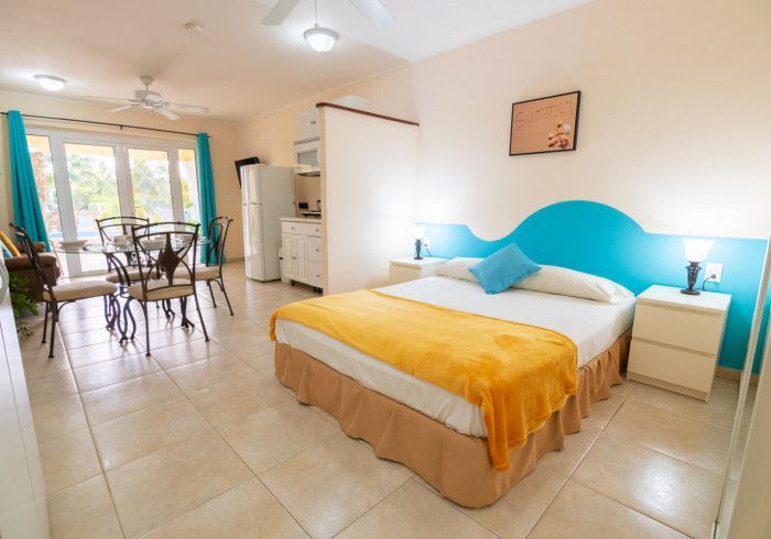 Bright bedroom with a yellow bedspread, blue headboard, tiled floor, and dining area by a window