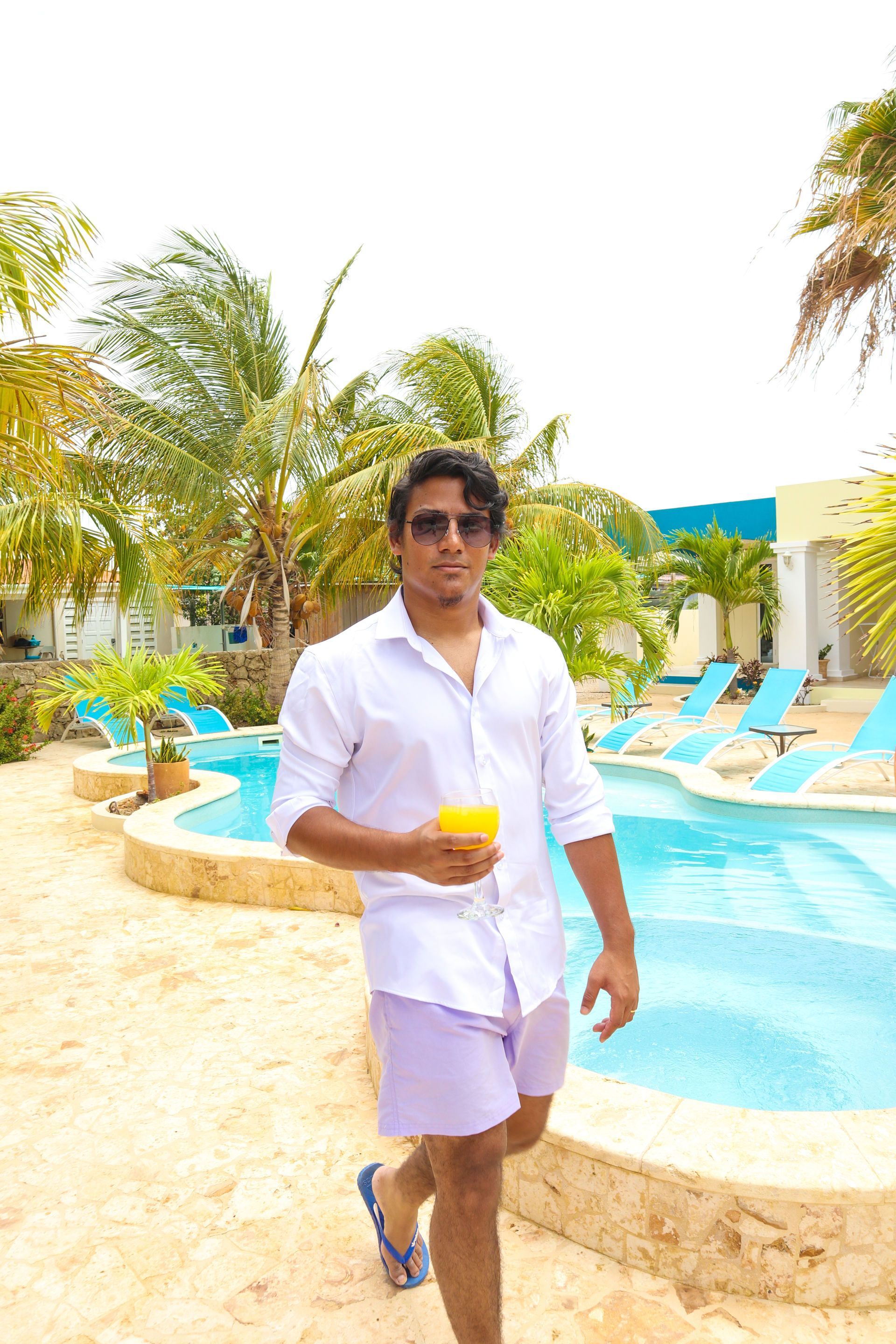 Man in white shirt and shorts holding a drink beside a resort pool with lounge chairs and palm trees