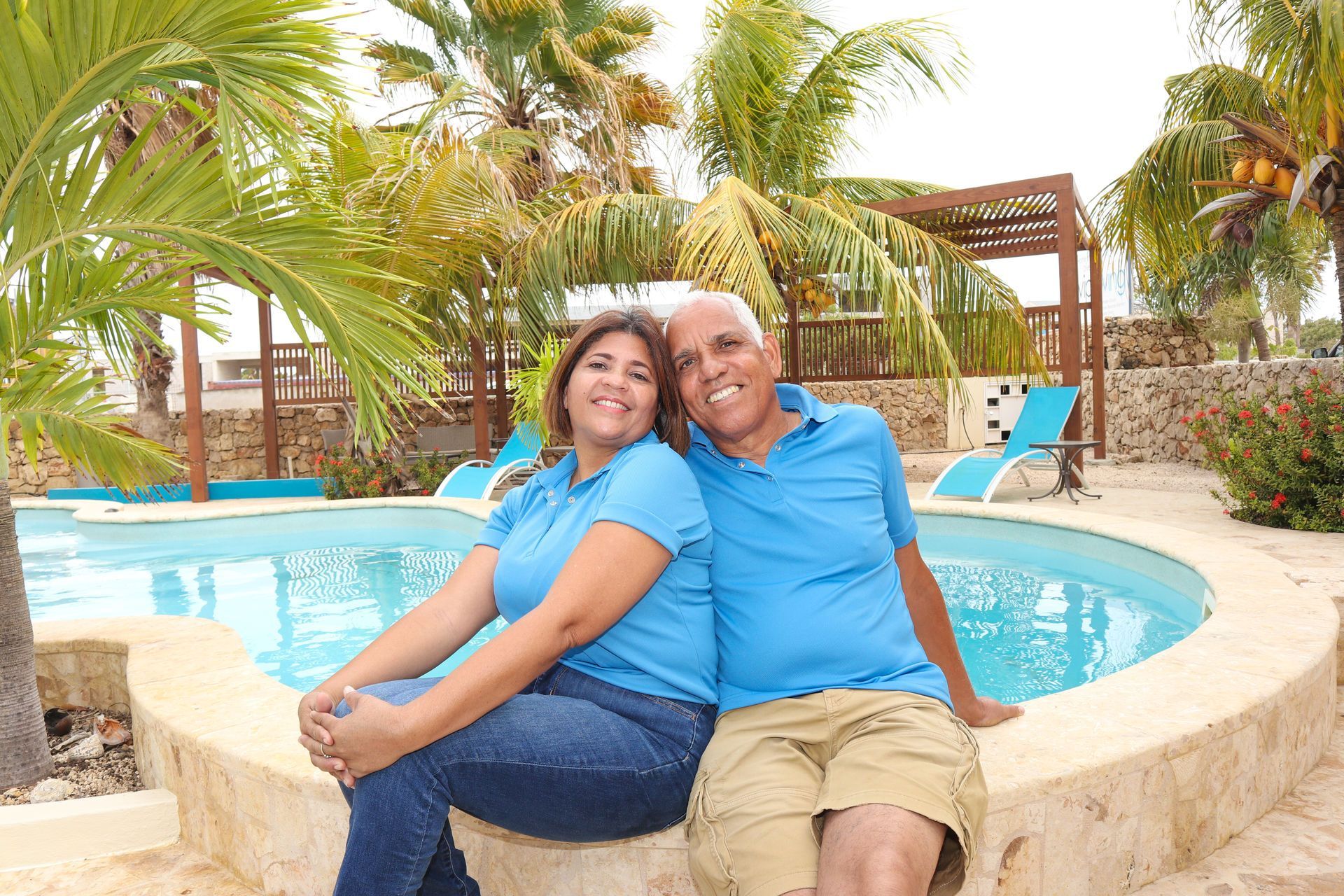 Couple in blue shirts, smiling by a pool with palm trees in sunny outdoor setting.