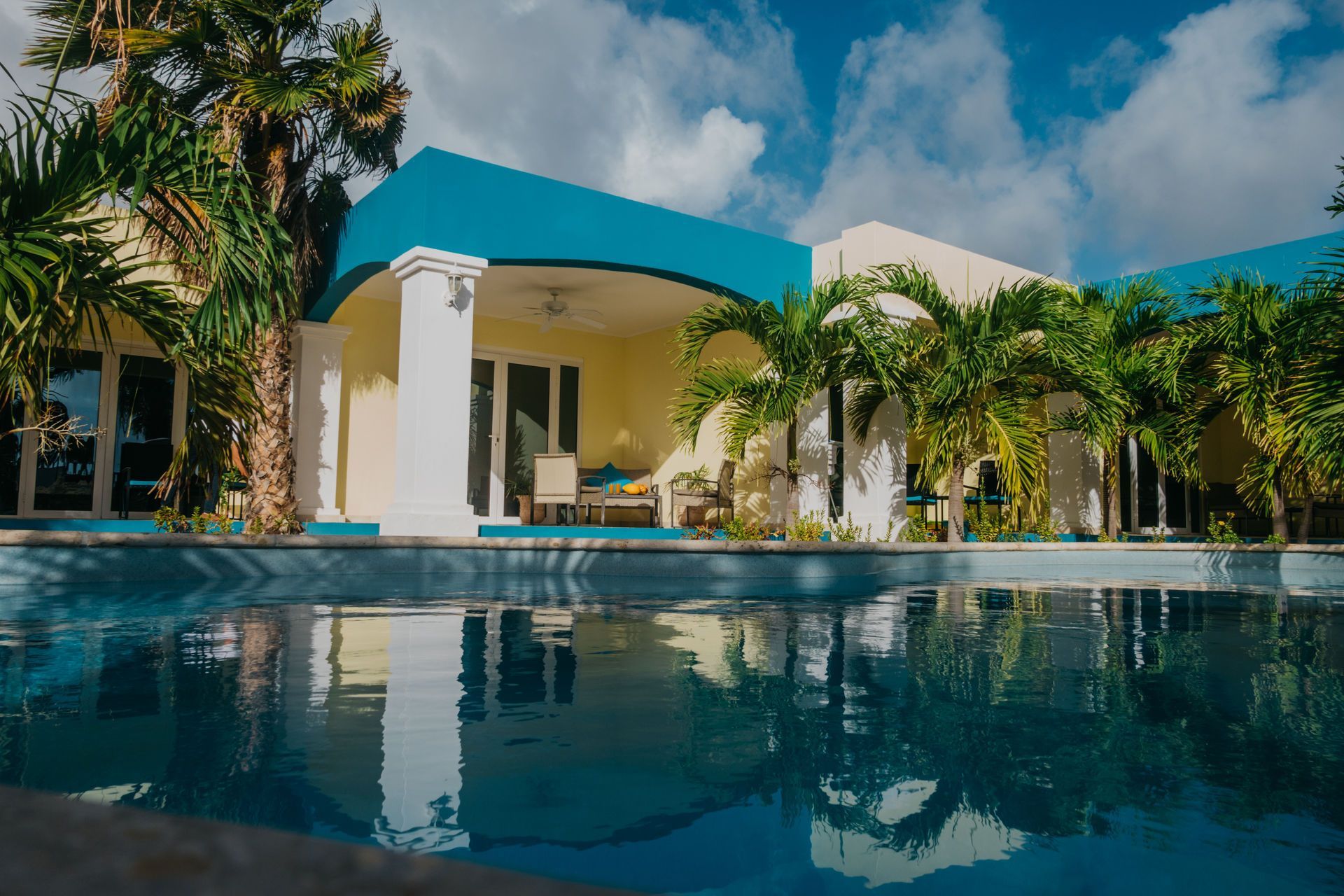 Swimming pool in front of a turquoise and yellow building with palm trees and a cloudy sky.