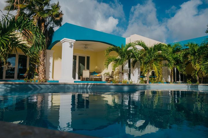 Modern villa with turquoise roof beside a swimming pool and palm trees under a cloudy sky
