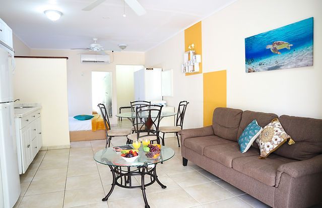 Bright living-dining room with brown sofa, glass table, white cabinets, and ocean artwork on the wall.