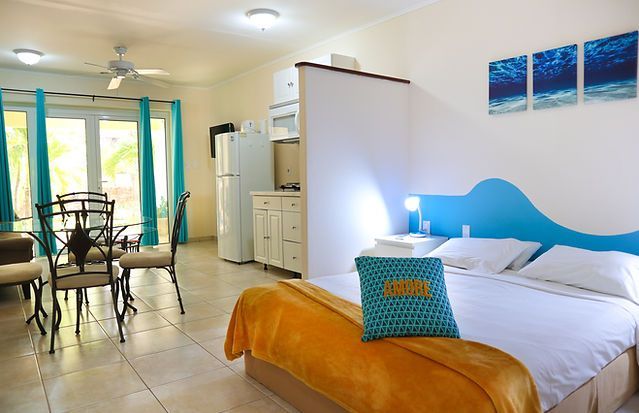 Bright hotel room with white bed, orange throw, blue headboard, dining table, and balcony doors.