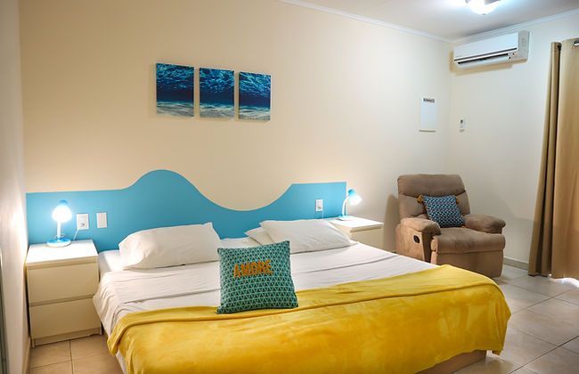 Bright hotel room with two twin beds, yellow bedspread, blue headboard, and a recliner by the window.