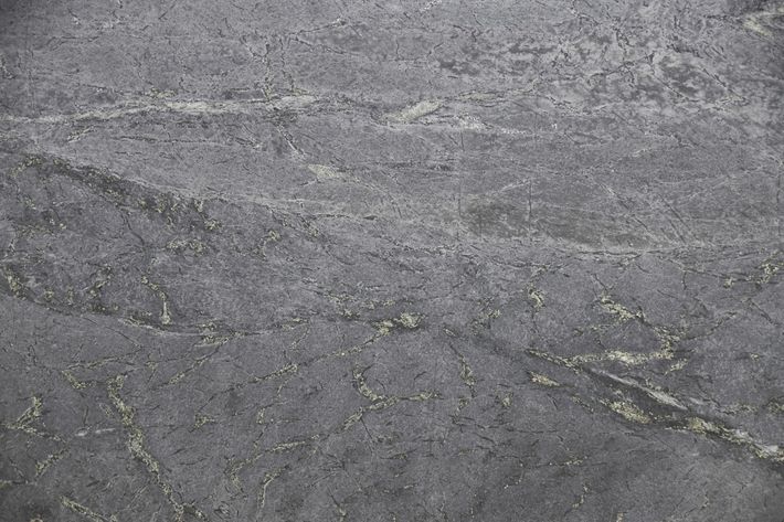 A close-up, top-down view of a textured dark gray stone surface with subtle white and green mineral veining throughout. A close-up, top-down view of a textured dark gray stone surface with subtle white and green mineral veining throughout.