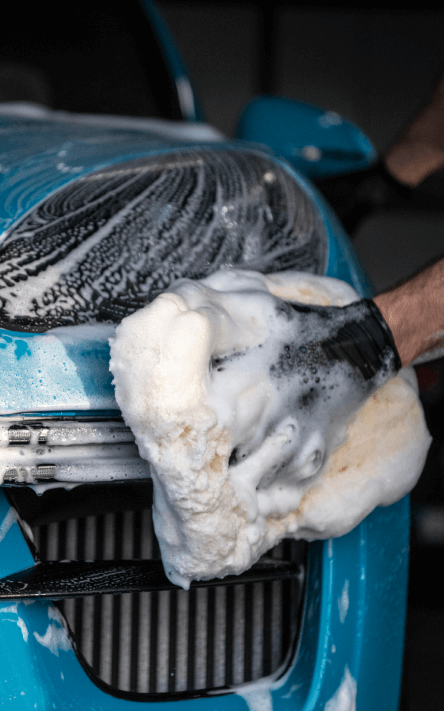 Soap foaming as someone scrubs the headlights of a blue car with a fluffy yellow sponge. 