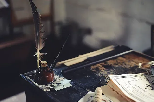 Quill pen in ink bottle on a cluttered desk with papers and a tray of wooden pens