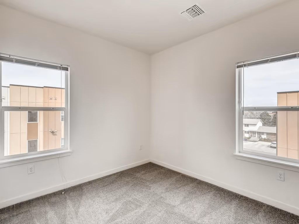An empty bedroom with white walls, carpeted floors, and two windows showing exterior buildings.