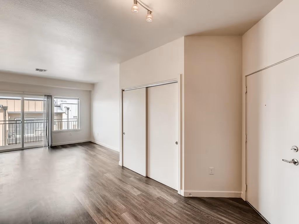 An empty apartment room with light beige walls, dark wood-look floors, sliding closet doors, and a glass balcony door.