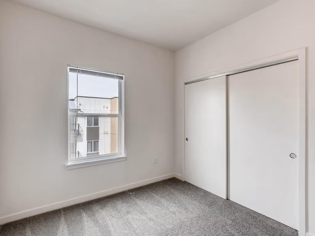 A sparsely furnished room with white walls, gray carpet, a single window, and a white sliding-door closet.