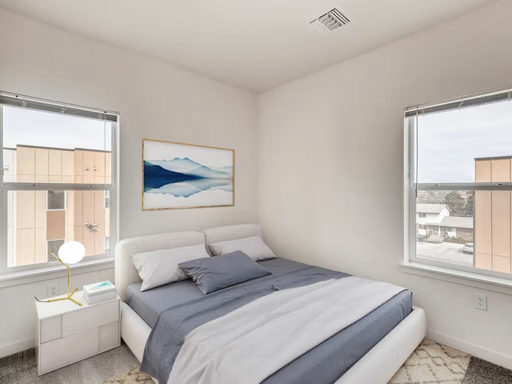 A bedroom with a queen bed featuring blue and white bedding, a white nightstand, and two windows with light beige walls.