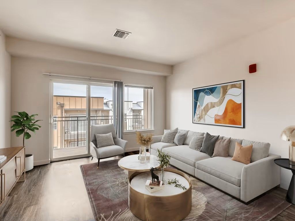 A modern, sunlit living room features a gray sofa with colorful pillows, nested coffee tables, and a patterned area rug.