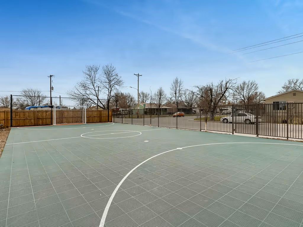 An outdoor green modular court with white boundary markings under a clear blue sky, bordered by wooden and metal fencing.