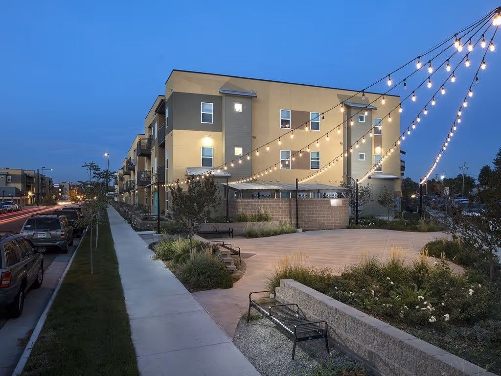 A multistory tan apartment building at dusk, with string lights overhead, a sidewalk, parked cars, and a landscaped plaza.