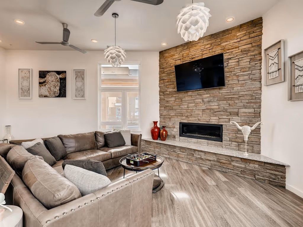 A modern living room with a large stone fireplace, brown sectional sofa, pendant lighting, and neutral decor.