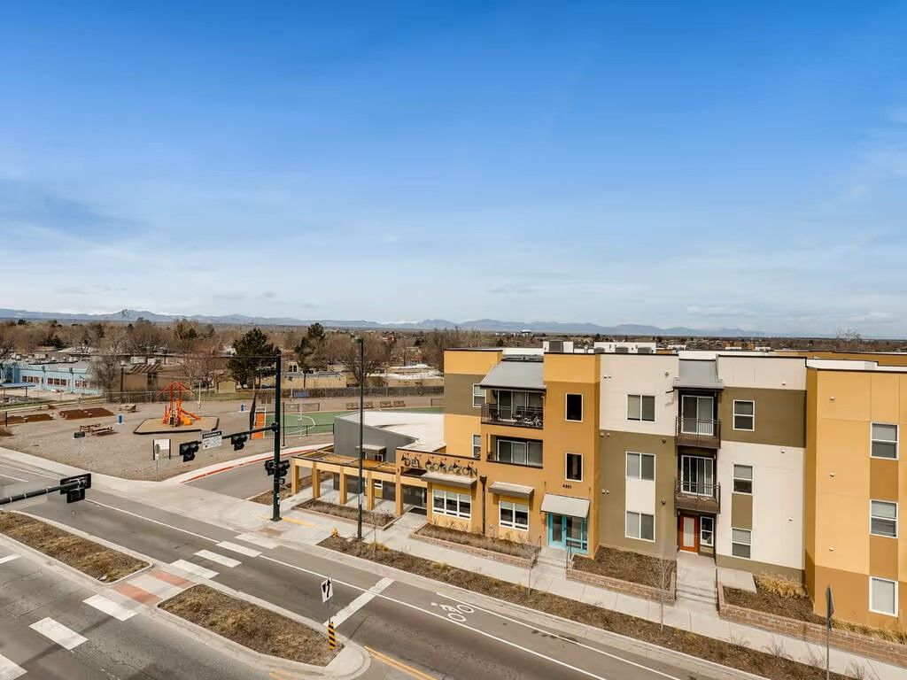 An aerial view of a multi-story apartment building with tan, brown, and white stucco walls, adjacent to a paved roadway.