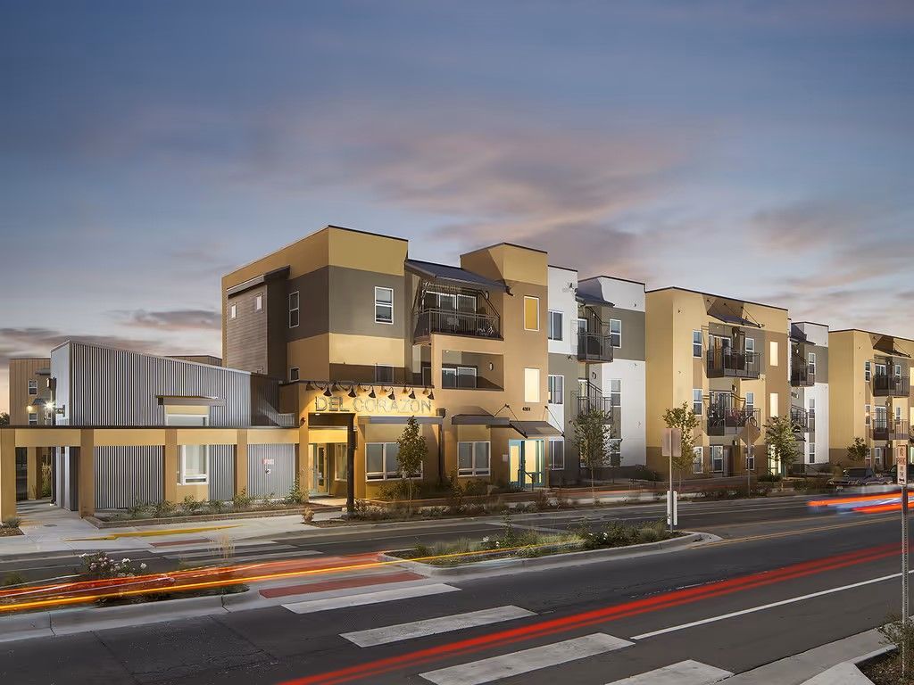 Modern apartment complex with tan and grey facade at twilight, featuring balconies and a pedestrian crosswalk.