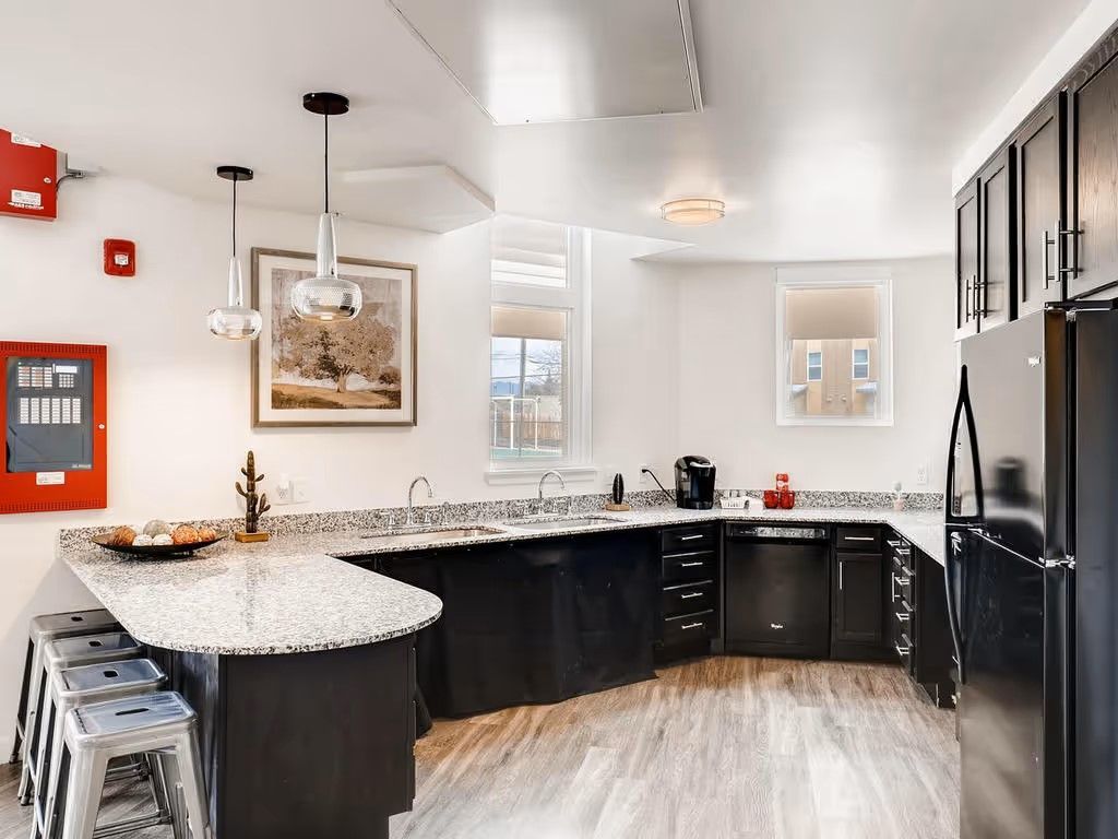 A kitchen with black cabinets, granite countertops, a bar with three metal stools, and a black refrigerator.
