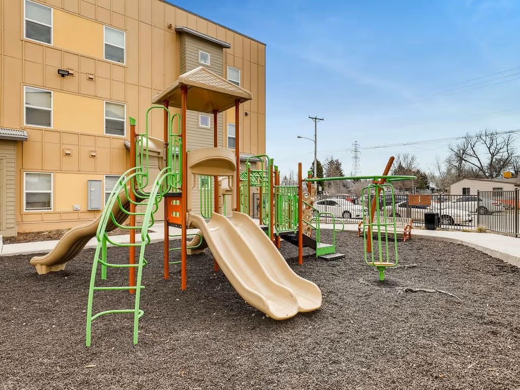 A tan and green modular playground set with two slides on a wood-chip surface next to a tan apartment building.