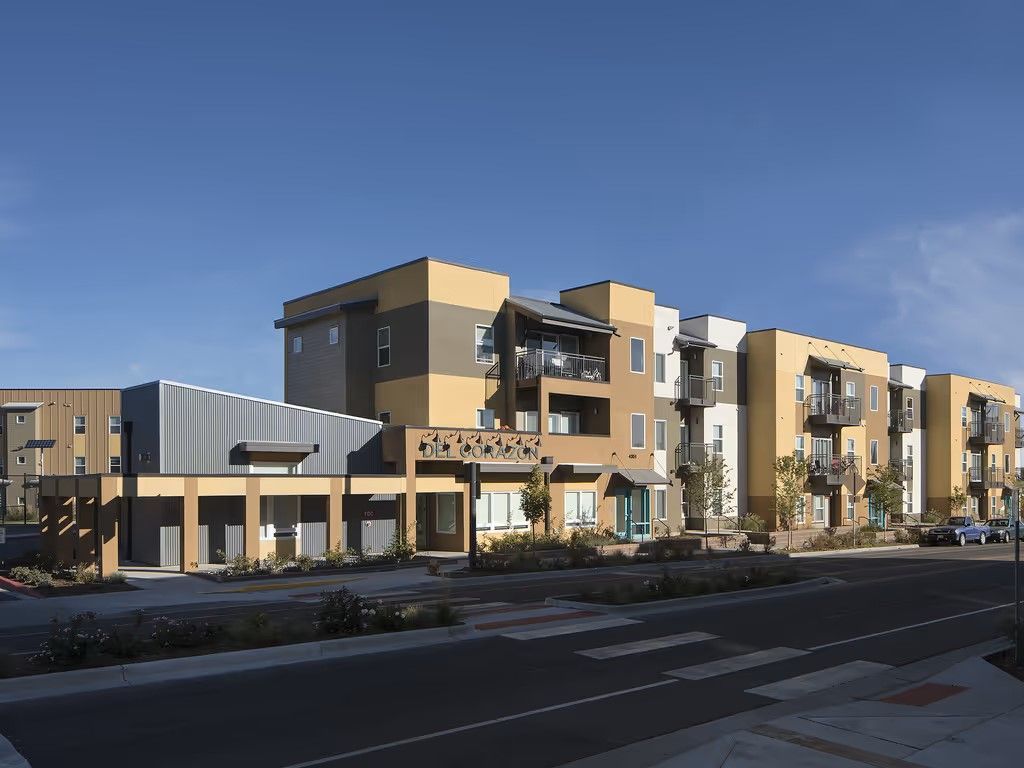 A multi-story modern apartment complex with tan and grey siding, featuring balconies, under a clear blue sky.