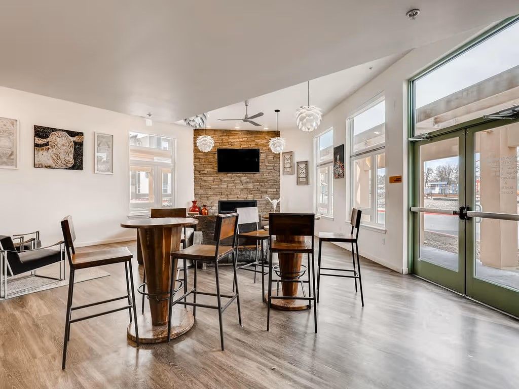 A bright, modern lounge featuring bar-height tables, stools, a stone fireplace with a wall-mounted TV, and glass entry doors.