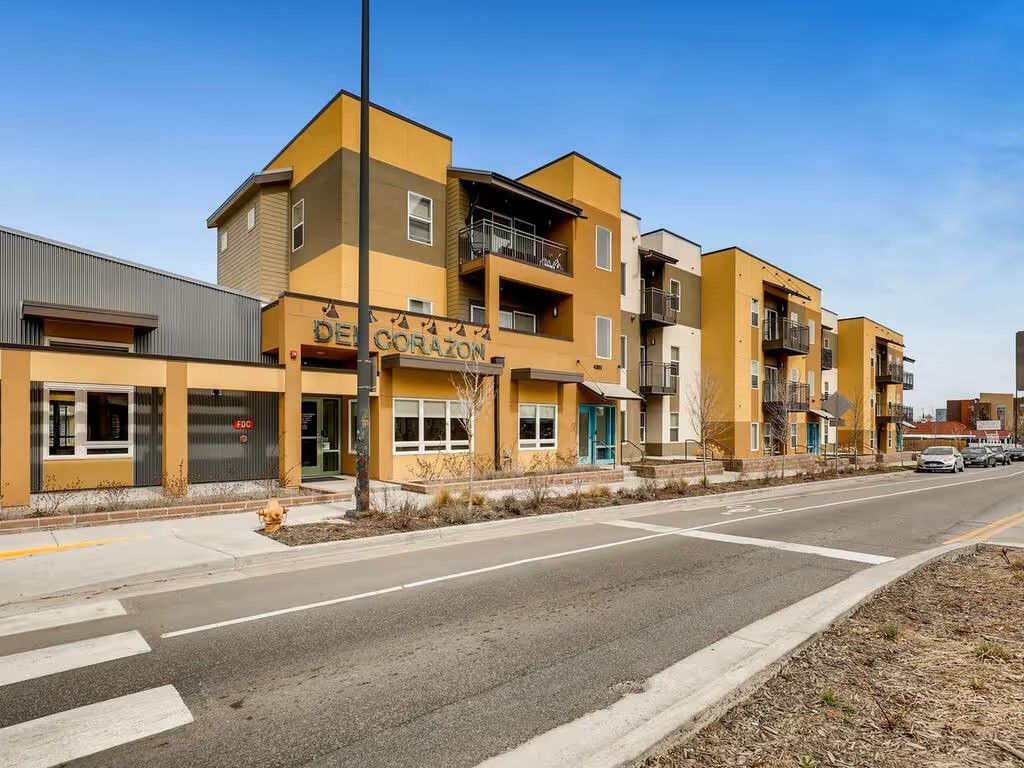 A multistory tan and brown apartment complex with balconies, set along a street with a crosswalk under a clear blue sky.