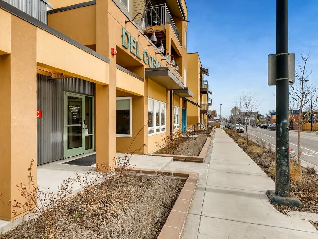 A tan apartment building exterior with green doors and a sidewalk leading toward a street on a clear day.