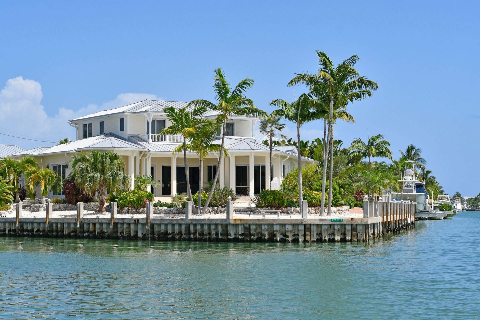 Tropical waterfront villa with white columns, palm trees, and a dock beside calm blue water under a sunny sky