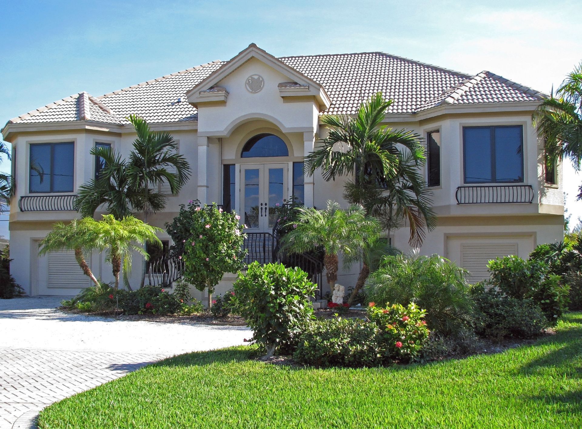 Large beige two-story house with a tiled roof, arched entry, and green lawn