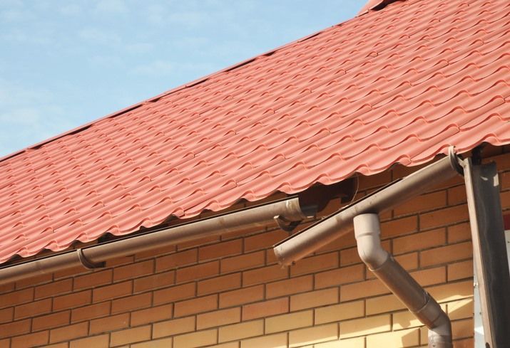 Red corrugated metal roof with brown gutters and a downspout, against a brick wall and blue sky.